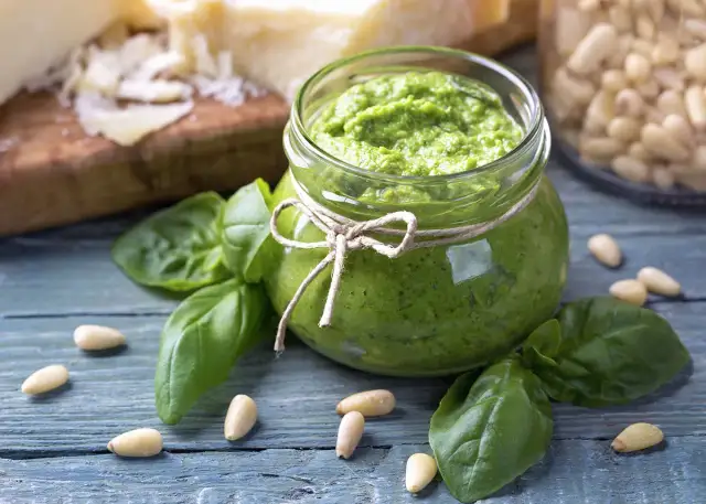 Słoik domowego pesto z bazylii, otoczony świeżymi liśćmi bazylii i orzeszkami pinii. Obok kawałki parmezanu. Idealne do makaronu.