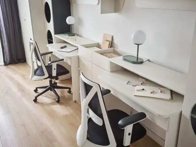 Mały pokój dla 3 dzieci z dwoma biurkami i krzesłami. Na biurkach lampki, zeszyty i kredki.