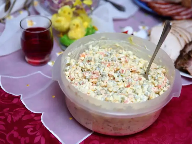 Sałatka jarzynowa w przezroczystej misce z łyżką, obok szklanka z napojem i wielkanocne ozdoby.