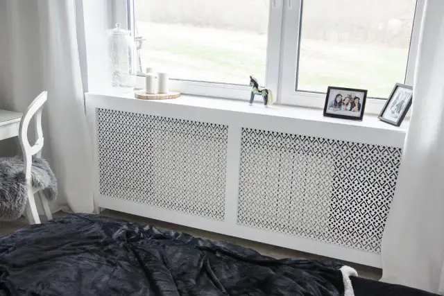 Elegancka osłona kaloryfera z ażurowym wzorem, która stanowi estetyczne rozwiązanie, jak zabudować kaloryfer. Na parapecie zdjęcia i ozdoby.