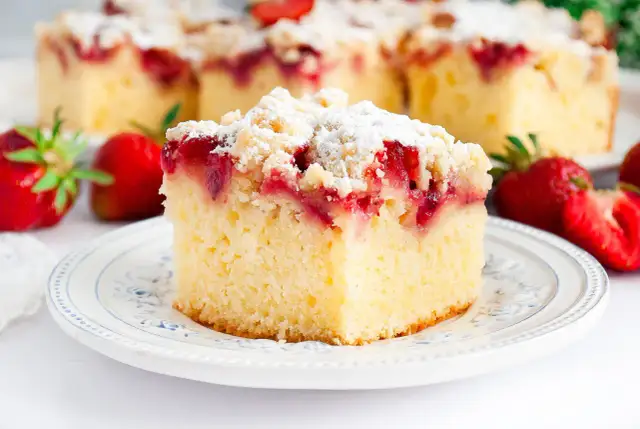 Puszyste ciasto jogurtowe z truskawkami i kruszonką, posypane cukrem pudrem. Obok świeże truskawki.