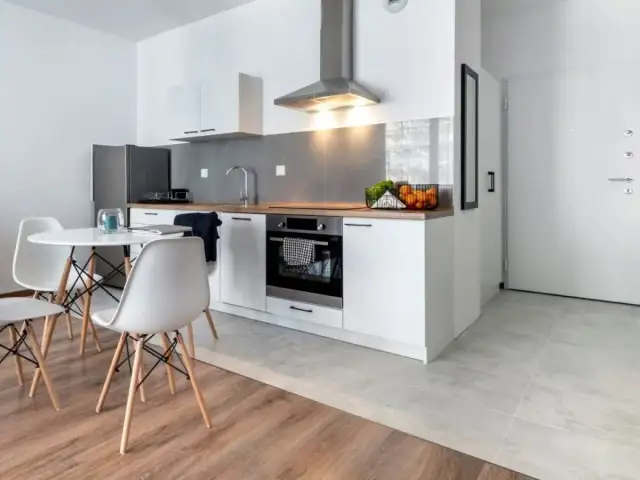 Nowoczesna kuchnia i jadalnia w małym mieszkaniu 35 m. Białe meble, okrągły stół z krzesłami, lodówka i piekarnik.