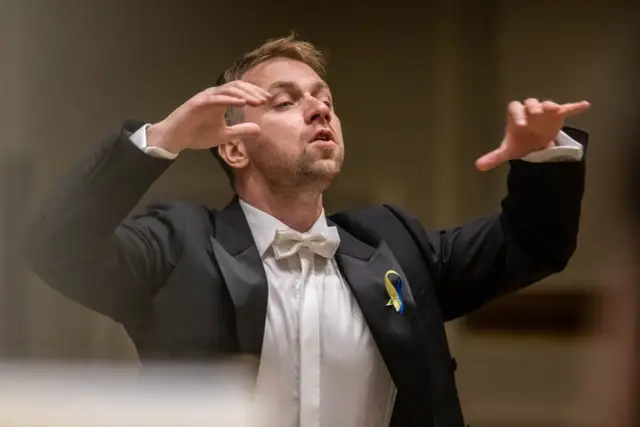 Dyrygent w garniturze z kokardką, prowadzący wykonanie Carmina Burana, z ukraińską wstążką na klapie.