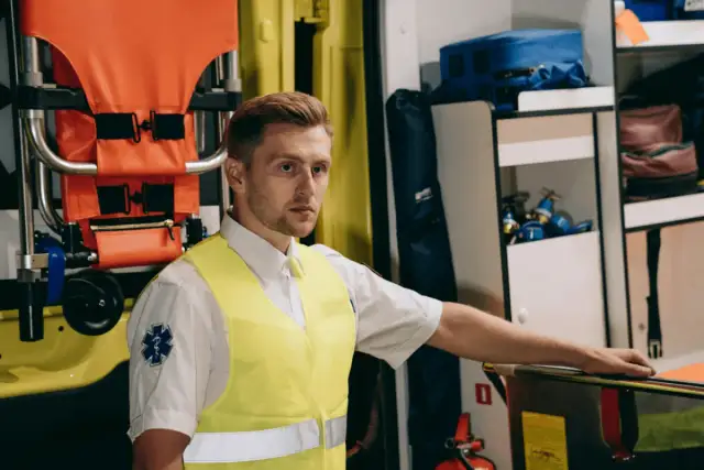 Ratownik medyczny w żółtej kamizelce stoi obok noszy w karetce.