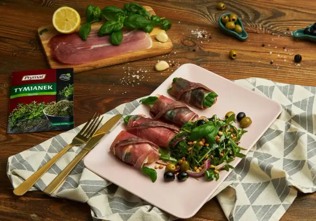 Roladki z szynką parmeńską i anchois, podane z rukolą, oliwkami i orzeszkami pinii. Obok talerza złote sztućce i opakowanie tymianku.