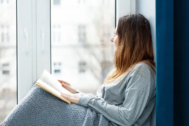 Kobieta w szarym swetrze czyta książkę, siedząc przy oknie i patrząc na zewnątrz.
