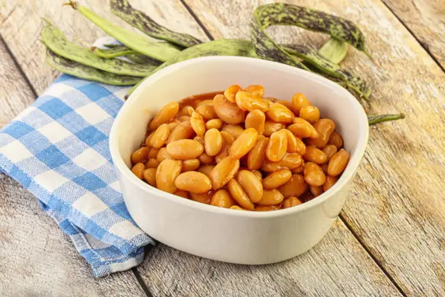 Miseczka fasolki w sosie na drewnianym tle, obok zielone fasolki i niebieska serwetka.