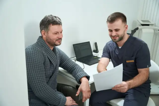 Lekarz w granatowym uniformie pokazuje pacjentowi w szarym szlafroku dokumenty.