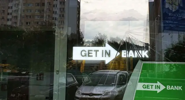 Czy lokaty w Getin Banku są bezpieczne? Znak "GET IN BANK" odbija się w szybie, obok samochodów i budynków.