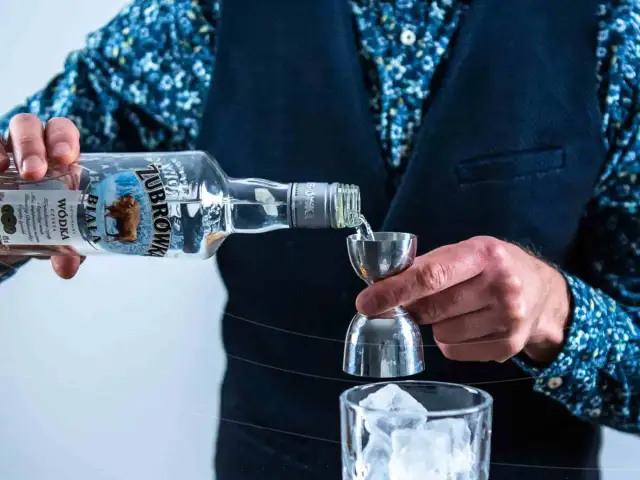 Barman nalewa wódkę Żubrówka Biała z butelki do miarki barmańskiej nad szklankę z lodem.
