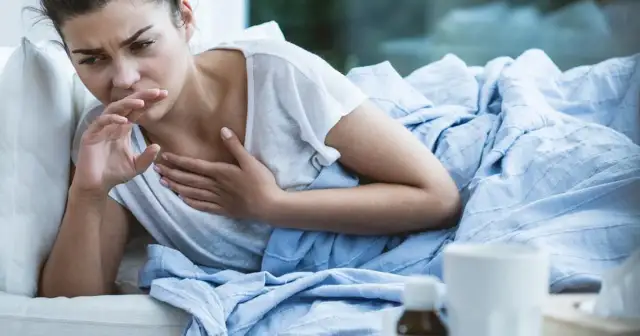 Kobieta w łóżku kaszle, zakrywając usta dłonią. Czuje się źle, może ma brązową flegmę.