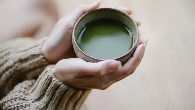 Dłonie w beżowym swetrze trzymają miseczkę z zielonym napojem. Czy matcha to zielona herbata? Tak, to sproszkowana zielona herbata.