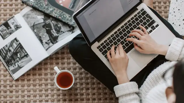 Kobieta pisze na laptopie, obok leży otwarta księga i filiżanka herbaty. Zastanawia się, ile kosztuje artykuł sponsorowany.