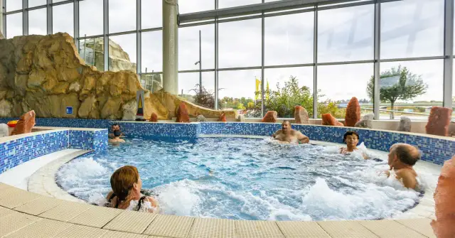 Ludzie relaksują się w basenie termalnym z hydromasażem, otoczonym skalnymi formacjami i dużymi oknami.