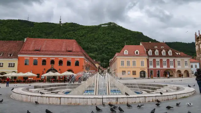 Fontanna na placu w Braszowie, z kolorowymi kamienicami i napisem "BRASOV" na wzgórzu. Wiele gołębi spaceruje wokół.