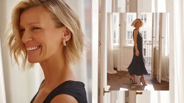 Uśmiechnięta kobieta w czarnej sukience i szpilkach stoi przy drzwiach balkonowych.
