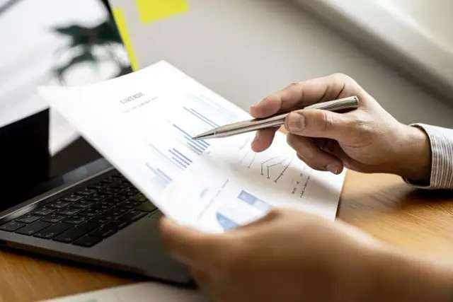 Analiza wykresów na papierze, wskazując długopisem. Skupienie na zarządzaniu kapitałem obrotowym i optymalizacji procesów.