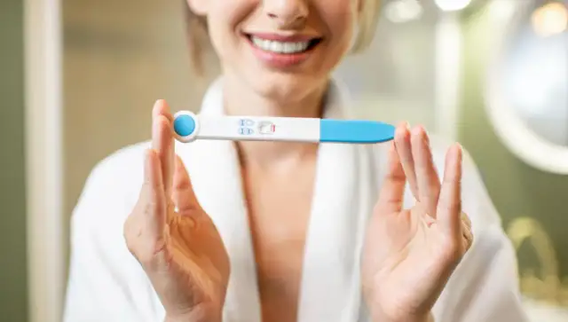Uśmiechnięta kobieta w szlafroku trzyma test ciążowy z dwoma kreskami. To moment, kiedy warto zastanowić się, kiedy zrobić test ciążowy.
