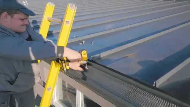 Dekarz przykręca siatkę na rynnie na dachu z blachy.