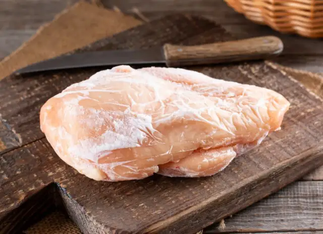 Zamrożona pierś kurczaka na drewnianej desce z nożem w tle.