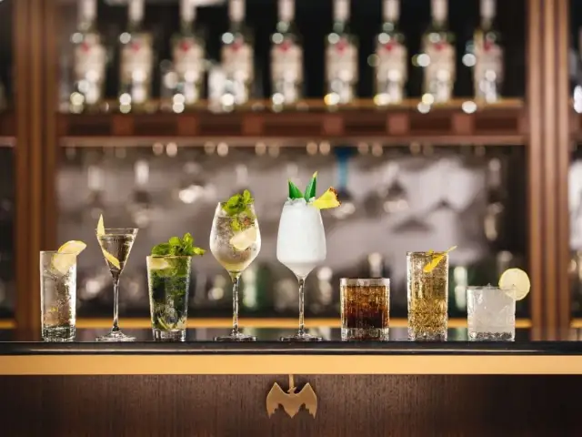 A row of seven colorful cocktails on a bar, with bottles in the background.