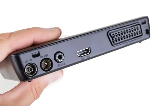 Dłoń trzyma czarny dekoder z portami RF IN, RF OUT, COAXIAL, HDMI OUT i SCART.