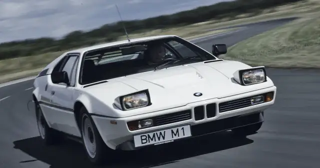 Białe BMW M1 na zakręcie drogi, z widocznym kierowcą i tablicą rejestracyjną.