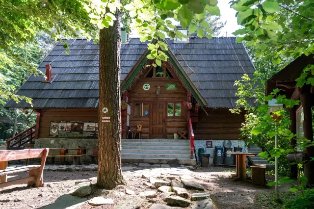 Drewniana bacówka PTTK w Bartnem, otoczona zielenią, zaprasza do odpoczynku. Na ścianie wisi tablica informacyjna.