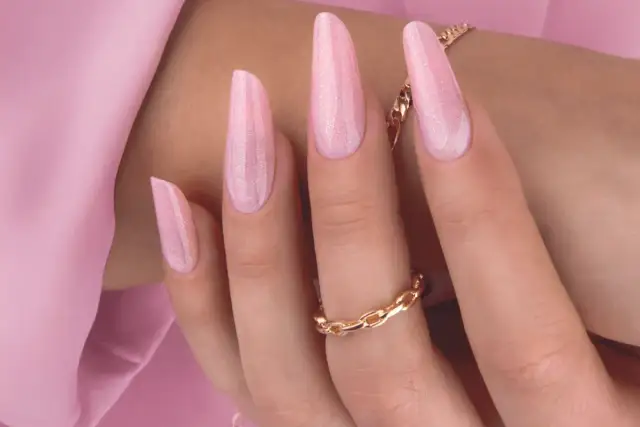 Delikatny, różowy manicure hybrydowy z subtelnymi drobinkami brokatu. Długie paznokcie w kształcie migdała, ozdobione złotą biżuterią.