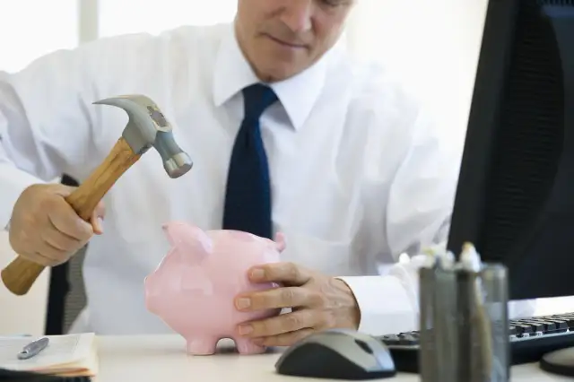 Mężczyzna w białej koszuli i krawacie zamierza rozbić skarbonkę młotkiem. Zastanawia się, ile trwa likwidacja lokaty, gdy pieniądze są w niebezpieczeństwie.