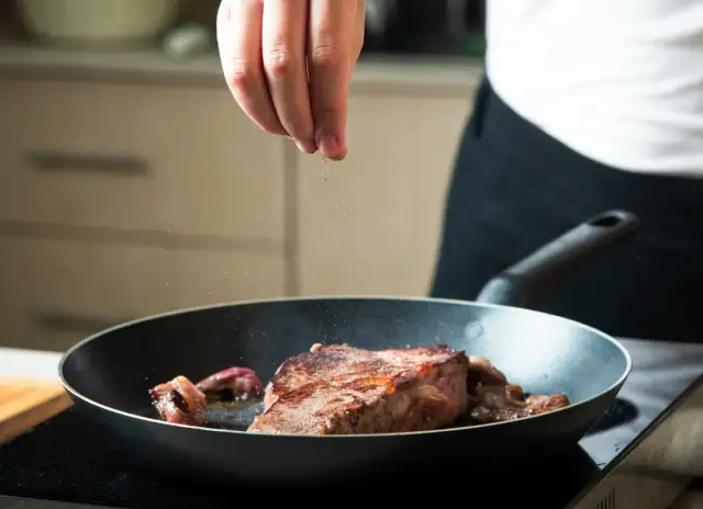 Ręka doprawia stek smażony na patelni.