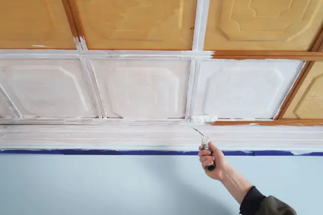 Ręka z wałkiem maluje kasetony na biało. Drewniane elementy sufitu zyskują nowy wygląd.