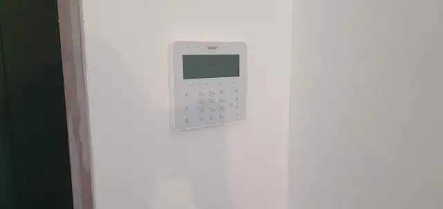 Biały panel alarmowy z klawiaturą numeryczną i ekranem na białej ścianie.