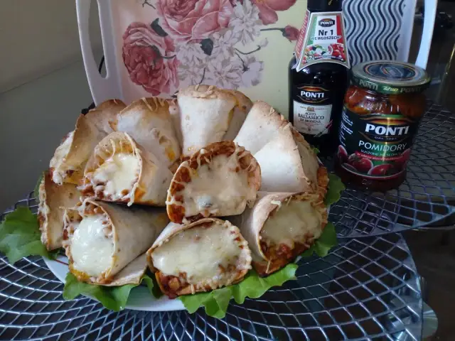 Pyszne rożki z tortilli z mięsem mielonym i ciągnącym się serem, podane na liściach sałaty. Obok butelka sosu balsamicznego i słoik pomidorów.