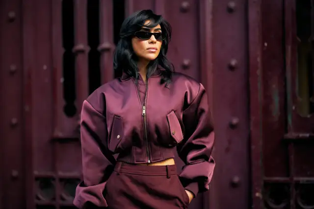 Kobieta w okularach przeciwsłonecznych i satynowej kurtce bomberce w odcieniu śliwki. Stylizacja nawiązuje do mody sprzed lat, z nutą retro.