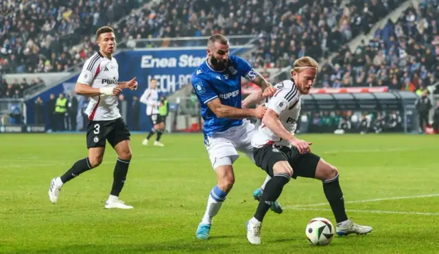 Zacięta walka o piłkę na boisku, kiedy pierwszy mecz ekstraklasy nabiera tempa. Dwóch zawodników walczy o kontrolę, podczas gdy trzeci biegnie w tle.