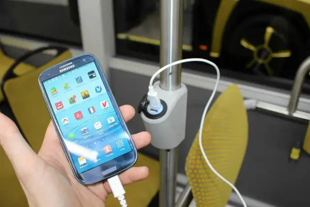 Dłoń trzyma smartfon podłączony do ładowarki USB zamontowanej na słupku w autokarze.