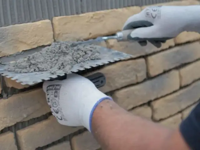 Ręce w rękawiczkach nakładają zaprawę na kamień dekoracyjny betonowy, przygotowując go do ułożenia na ścianie.