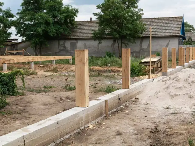 Budowa fundamentu pod ogrodzenie: betonowy murek, drewniane słupki i piasek.