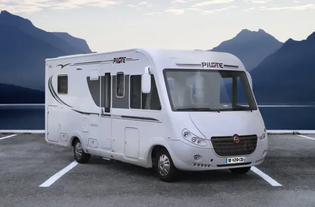 Biały kamper z prysznicem zaparkowany nad jeziorem, w tle góry.