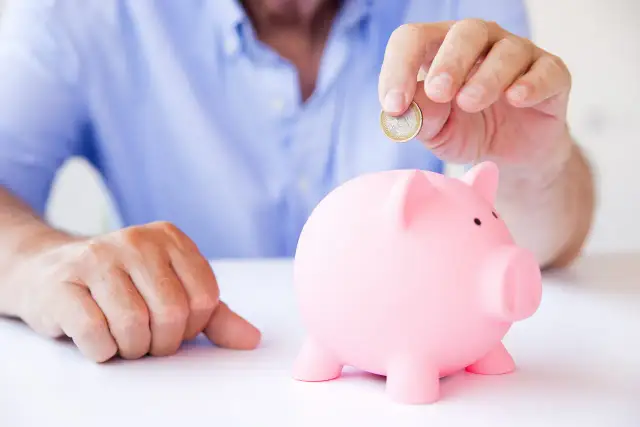 Mężczyzna wrzuca monetę do różowej świnki-skarbonki, zastanawiając się, czy lepsze będą lokaty czy konto oszczędnościowe.
