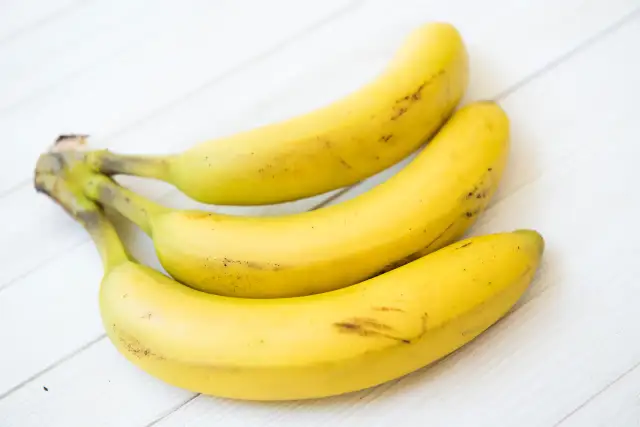 Trzy dojrzałe banany leżą na białym, drewnianym blacie. Czy banany tuczą? Zależy od ilości i sposobu ich spożycia.