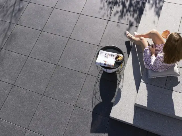 Kobieta siedzi na schodach tarasowych z kawą i gazetą, na tle szarej kostki brukowej.