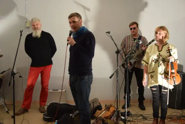 Orkiestra Świętego Mikołaja przygotowuje się do występu. Wokalista z mikrofonem, gitarzysta, skrzypaczka i brodaty mężczyzna w czerwonych spodniach.