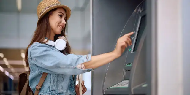 Uśmiechnięta kobieta w kapeluszu i dżinsowej kurtce dokonuje wypłaty z bankomatu za granicą.