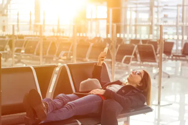 Kobieta leży na lotnisku, sprawdzając telefon. Czeka na lot, zastanawiając się, ile czasu trwa odprawa na lotnisku.