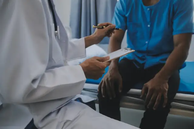 Lekarz w białym fartuchu rozmawia z pacjentem, trzymając notatnik i długopis.