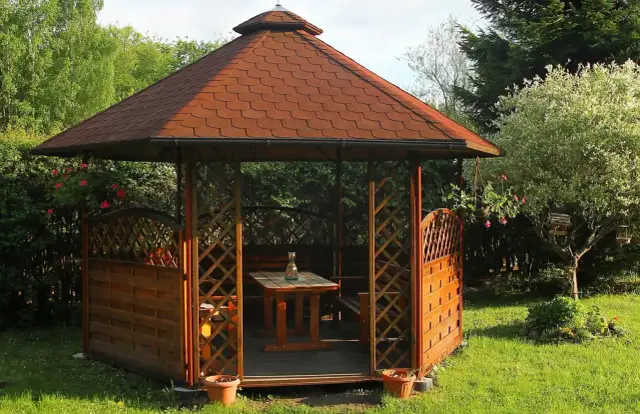 Drewniana altana ogrodowa z czerwoną, łuskowatą papą na dachu, otoczona zielenią.