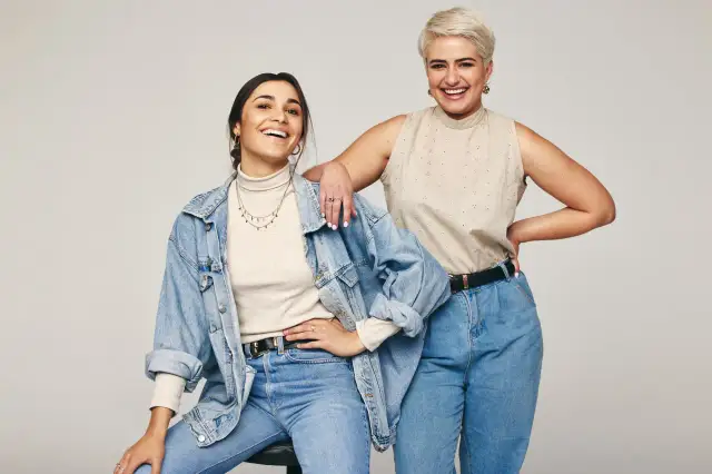 Dwie uśmiechnięte kobiety w jeansach. Jedna w beżowym golfie i jeansowej kurtce, druga w beżowej bluzce bez rękawów. Inspiracja: jaka bluzka do jeansów.