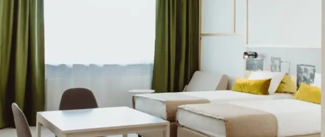 Pokój hotelowy z dwoma łóżkami, stołem i krzesłami, zielonymi zasłonami.